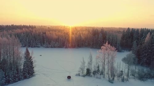 Aerial drone forward moving shot over a frozen lake beside rectangular cottage and barrel sauna on a