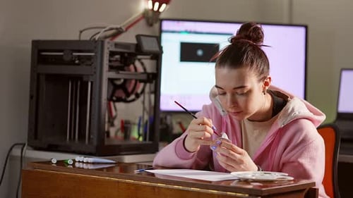 Young Adult Woman Sits at Table Painting and Decorating Small 3D Printed Figure