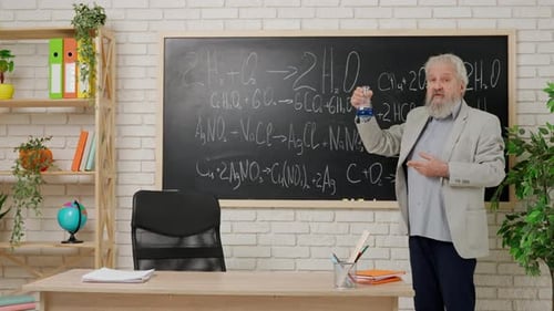 Aged Man College Professor Standing at Desk in Classroom in Front of Chalkboard Explains Chemistry