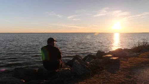 Man in baseball cap sitting down on sea shore rocks to watch sunset on horizon, Lion's Bridge, Newpo