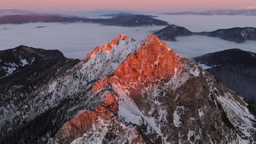 Aerial View of Snowy Rocky Peak Illuminated By Sunset Light with Fog Draping the Valley Below