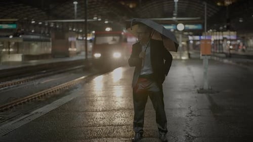 A Businessman Waiting on a Rainy Train Platform at Night in the City Surrounded By Urban Scenery