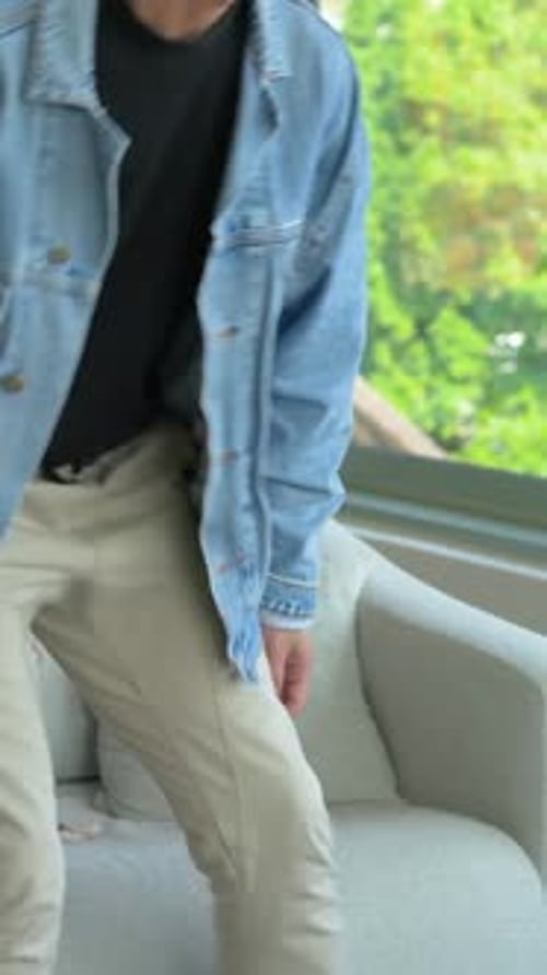 Vertical video: Reaching for mug, denim-jacket man moving to sofa at window, adjusting headphones