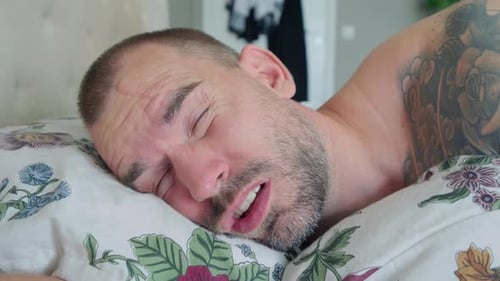 Sleeping Man Being Awakened By An Alarm Clock Inside His Bedroom. Close Up