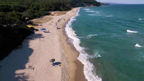 Aerial footage of a scenic sandy beach with waves gently hitting the shore, people relaxing under um