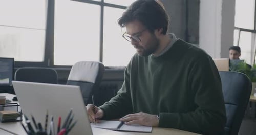 Young Businessman Using Laptop Typing and Writing in Notebook Working at Desk in Coworking Office
