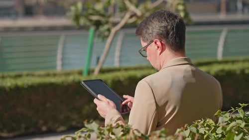 Busy Middle Aged Businessman Workin on Tablet Sitting on Bench in City Park