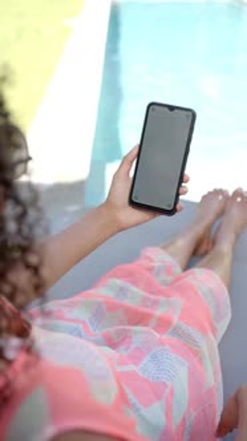 Vertical video: Relaxing by pool, person holding smartphone and lounging in colorful dress