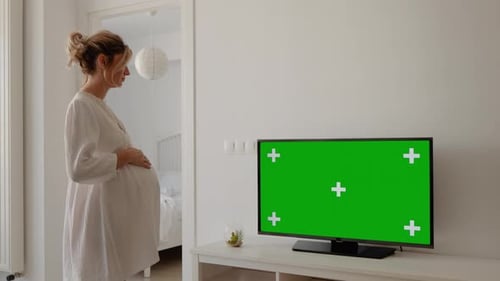 Pregnant woman coming from bedroom watching TV with green screen in living room.