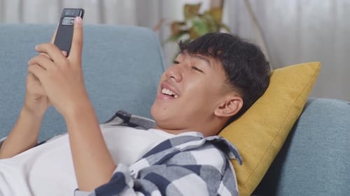 Close Up Of Asian Teen Boy Enjoys Playing Smartphone While Lying On Sofa In The Living Room