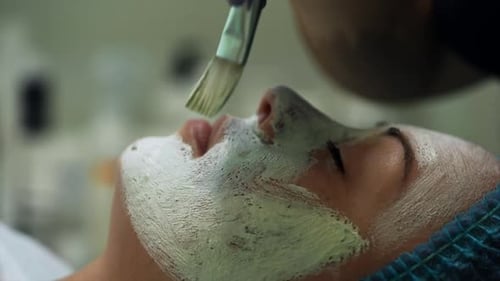 La esteticista pone una máscara blanca en el rostro de la clienta.