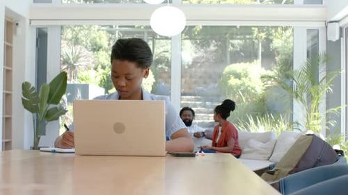 Adolescente africaine dactylographiant et écrivant sur une longue table en bois, faisant une pause pendant que les parents discutent