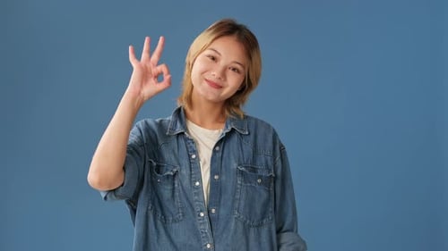 Smiling young woman looking at camera showing ok gesture isolated on blue background in studio