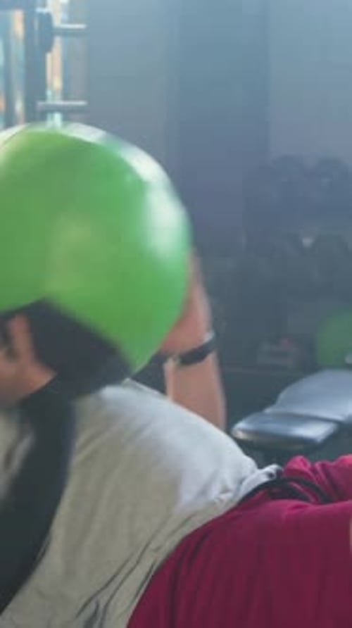 Vertical video: Bracing feet man in tee and shorts picking medicine ball on gym bench toning core