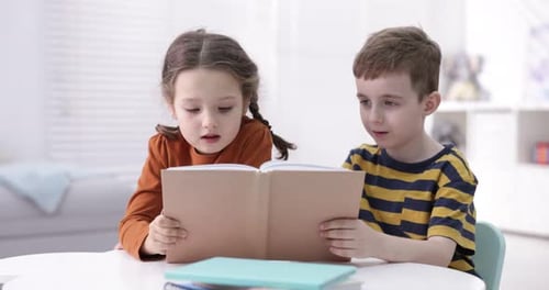 Cute little kids reading book at white table indoors