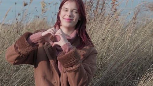 Smiling teen girl forms a heart shape with her hands, looking at the camera in a grassy field.