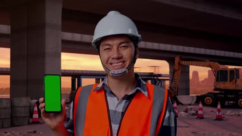 Close Up Of Asian Male Engineer Smiling And Showing Green Screen Smartphone At Building Site