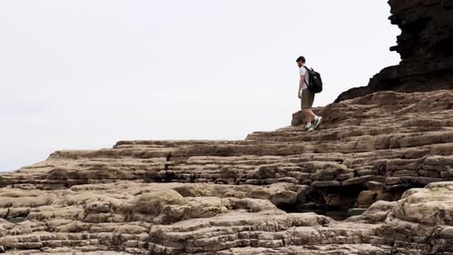 A young man walking across some large rocks on a beach.