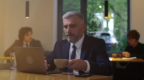 Mid-Aged Businessman Drinking Coffee and Working on Laptop in Cafe