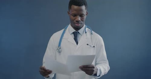 Portrait of African American Doctor in White Gown Reading Medical Records Holding Paper on Gray