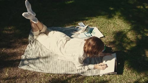 An Adult Relaxed Woman Lying on the Blanket in the Blooming Park and Drawing in Her Sketchbook