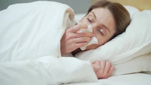 Femme blonde malade se mouchant, éternuant et toussant allongée sur le lit chaud de la chambre