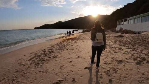 Woman walks along beach as sun sets behind the mountains