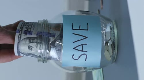 Poner billetes de dólar a mano en un tarro de cristal con la inscripción Guardar. Vídeo vertical