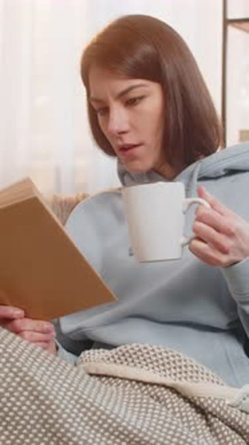 Smiling Young Woman Reading Interesting Book While Having Cup of Hot Chocolate on Couch at Home