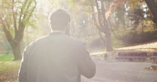 Vue arrière d'un jeune homme caucasien en veste marchant dans un parc par une journée ensoleillée d'automne et parlant sur mobile