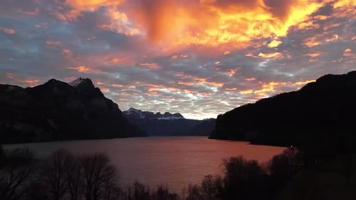 Dramático pôr do sol sobre um lago calmo na montanha com silhuetas escuras e nuvens brilhantes no céu.