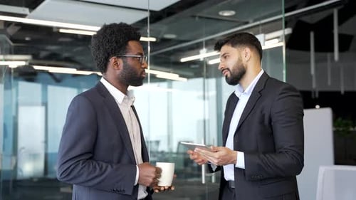 Two businessmen standing in modern office lobby holding coffee cups, discussing work matters.