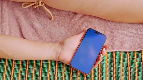 A Woman Holds a Smartphone Mockup With Tracking Marks While Lying on a Beach Towel as Warm Tropical