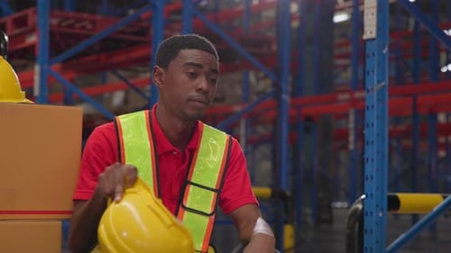 A tired black male warehouse worker sits in a paper warehouse, removes his helmet, and wipes sweat f
