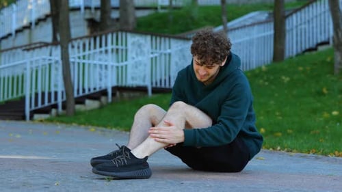 Sportsman Sitting on Ground and Stretching His Knee Leg Muscles in Green Park Before Outdoor