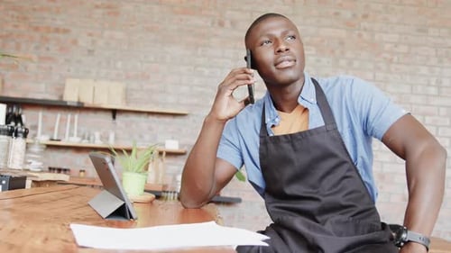 African american male barista using tablet and talking on smartphone at coffee shop, slow motion