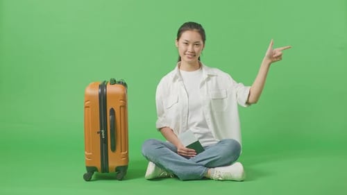 Asian Female Traveler With Luggage And Passport Smiling And Pointing To Side In The Green Screen