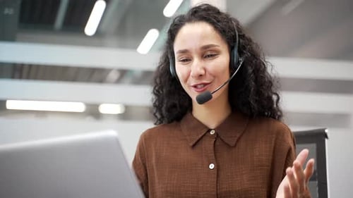 Smiling businesswoman in a wireless headset talking on a video call using a laptop sitting in office