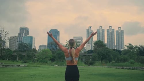 Strong blonde female doing sun greeting yoga pose in city park with high rise buildings in backdrop