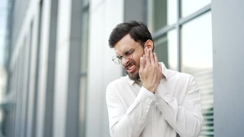 Upset businessman suffers from ear pain while standing on street near an office building.