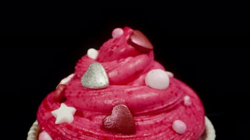 A cupcake in red packaging with pink frosting and silver hearts on it. Dolly slider, close up.