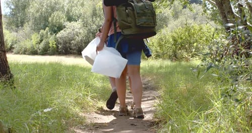 Una persona camina por un sendero de tierra con una mochila grande y un recipiente blanco, recolectando basura
