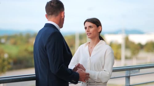 Two businesspeople stand outside next to a railing talking about business