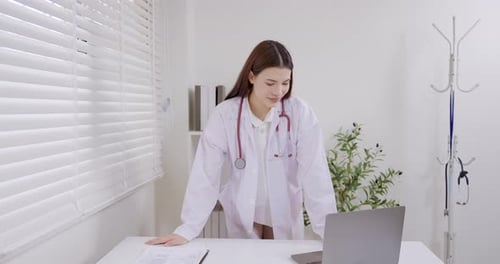 Asian female doctor in clinic reviewing documents and laptop on desk, showing dedication and