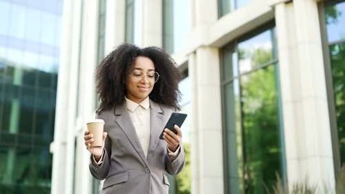 Black woman in business suit walks holding phone and coffee cup outside modern office building.