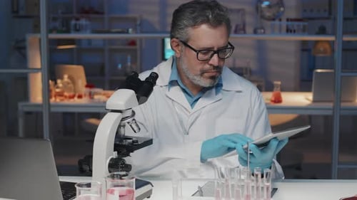 Mature Male Clinician in Lab Coat Using Tablet and Microscope