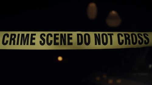 Close-up of crime scene tape. Flashing emergency lights and evidence markers can be seen in the back