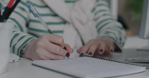 Closeup of Female Hand Writing in Notebook Pointing at Computer Screen at Desk in Office
