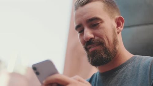 Close-up of young adult male with beard using cellphone from outside