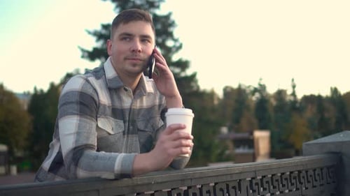 A Young Man Speaks on the Phone with a Glass of Coffee in His Hand A Man with a Phone Near the Fence